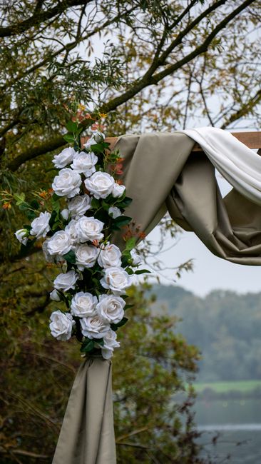 A floral arrangement featuring a cascading cluster of white roses and small pink blossoms intertwined with green foliage, attached to a rustic wooden arch draped with beige and white fabric. The bouqu